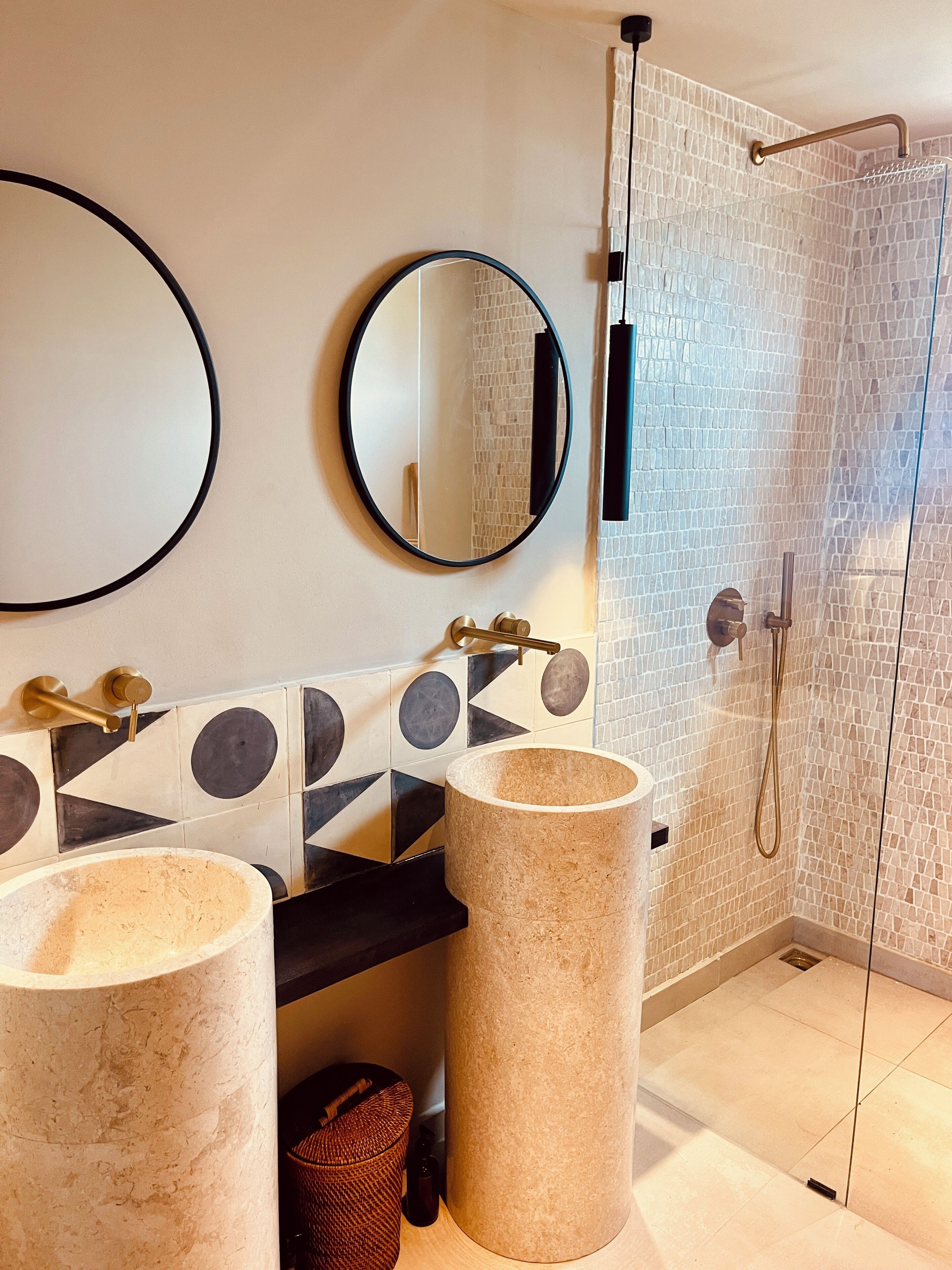 A modern bathroom with two large marble sinks, a round mirror, and a glass shower enclosure.