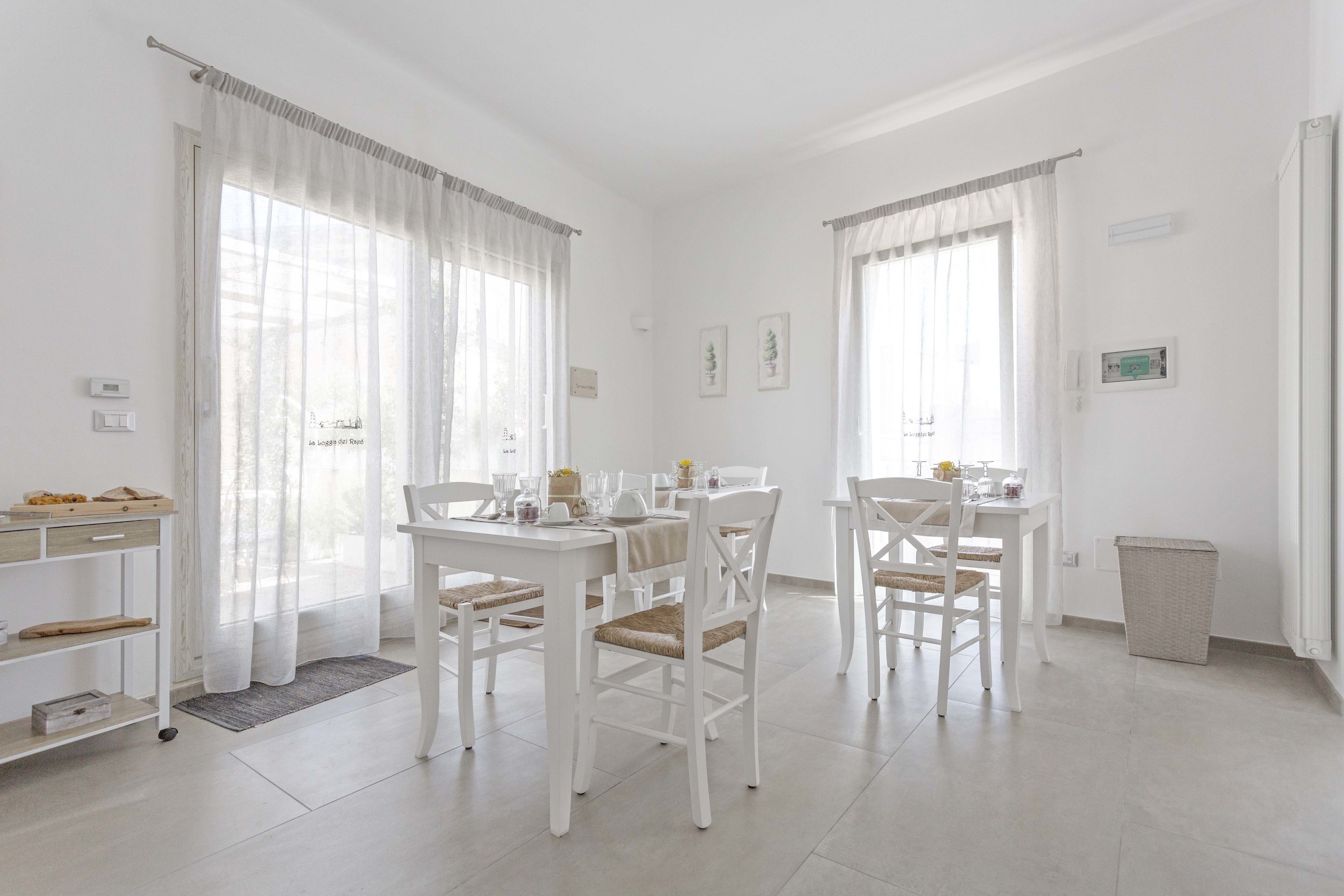 A bright, white dining area with two tables and chairs, a small shelf with items, and a trash bin.