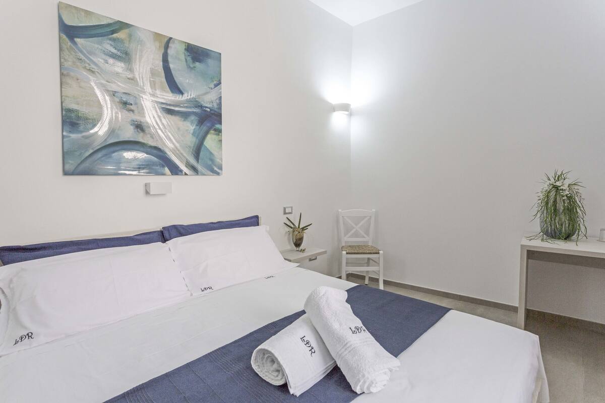 A hotel room with a bed, a painting, a desk, a chair, and a plant.