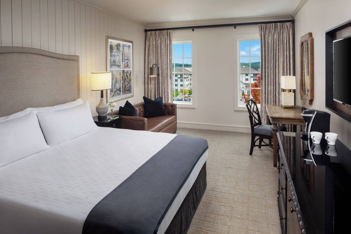 A hotel room with a large bed, a desk with a chair, a coffee machine, and a TV. There are two windows with curtains, a painting on the wall, and a lamp on the bedside table.
