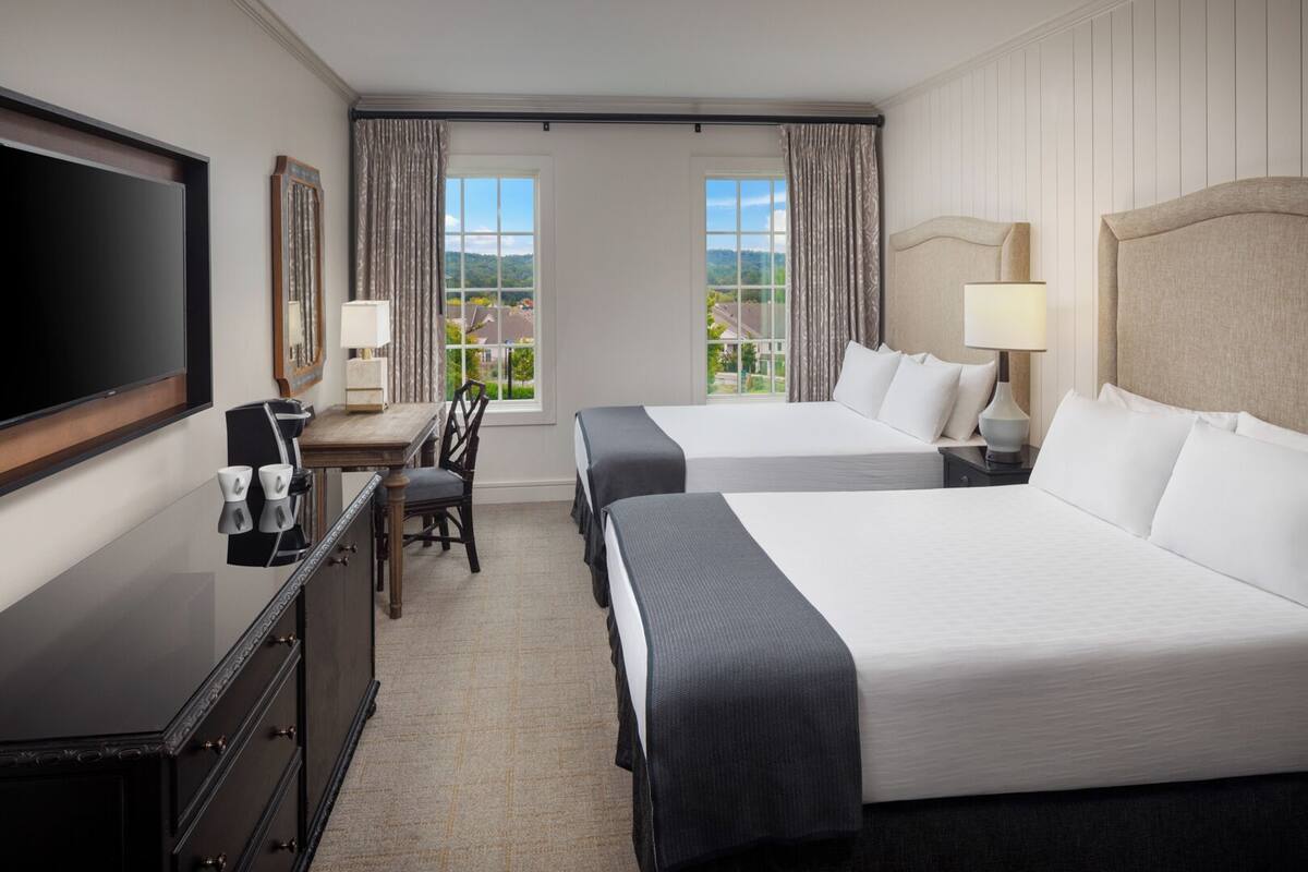 A hotel room with two beds, a desk, a TV, and a window with a view.