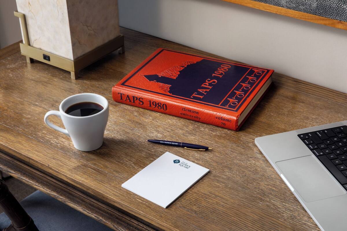A wooden desk with a laptop, a coffee cup, a pen, a notebook, and a book titled "TAPS 1980".