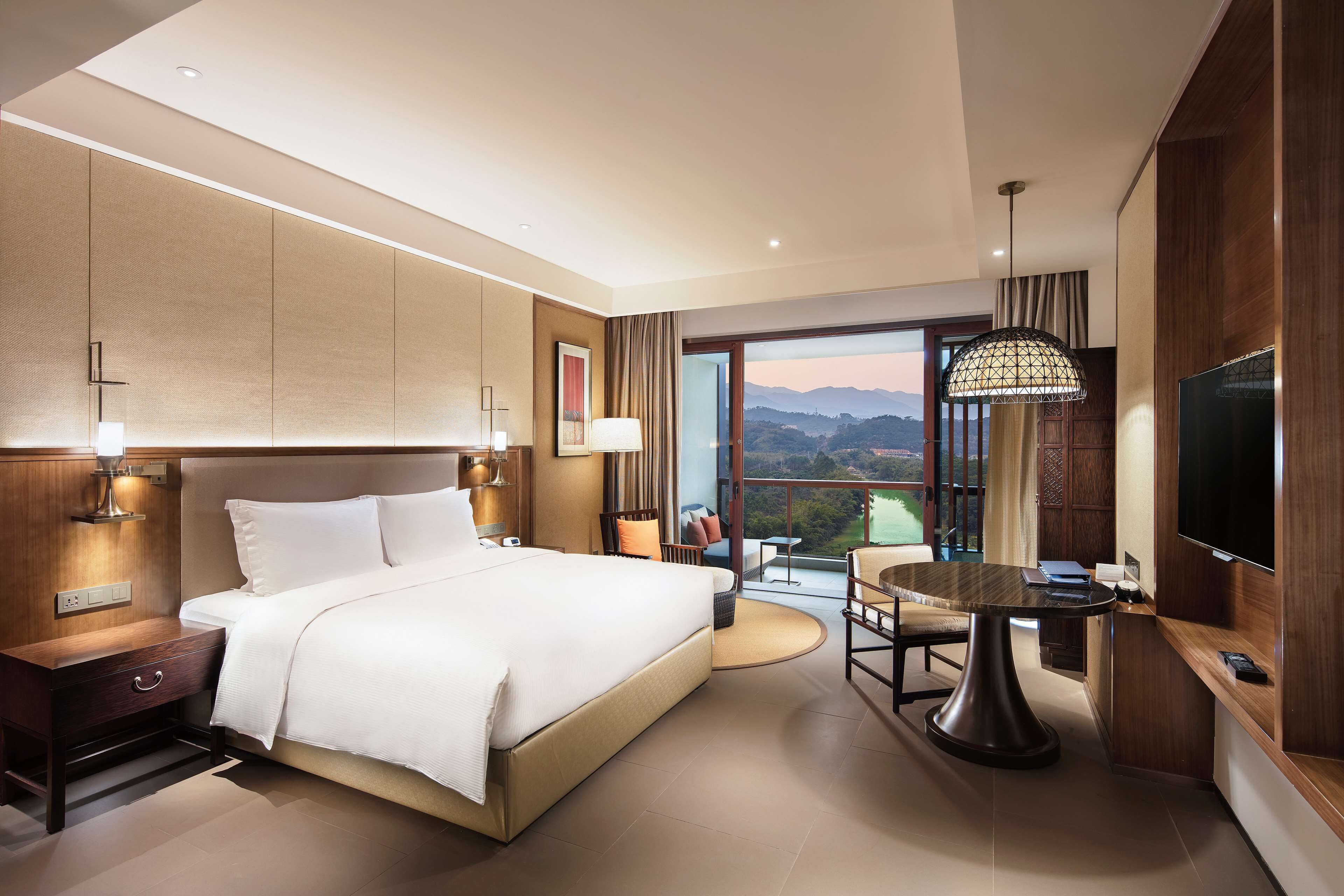 Habitación de hotel moderna con una cama grande, una mesa de comedor, un televisor y un balcón con vistas a la montaña.
