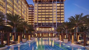 Een meerlaags hotel met een zwembad ervoor, omgeven door palmbomen en ligbedden.