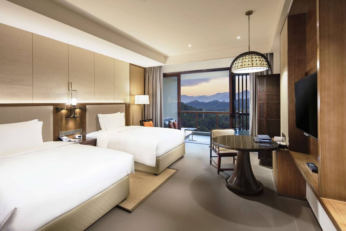 A hotel room with two beds, a desk, a TV, and a balcony with a view.