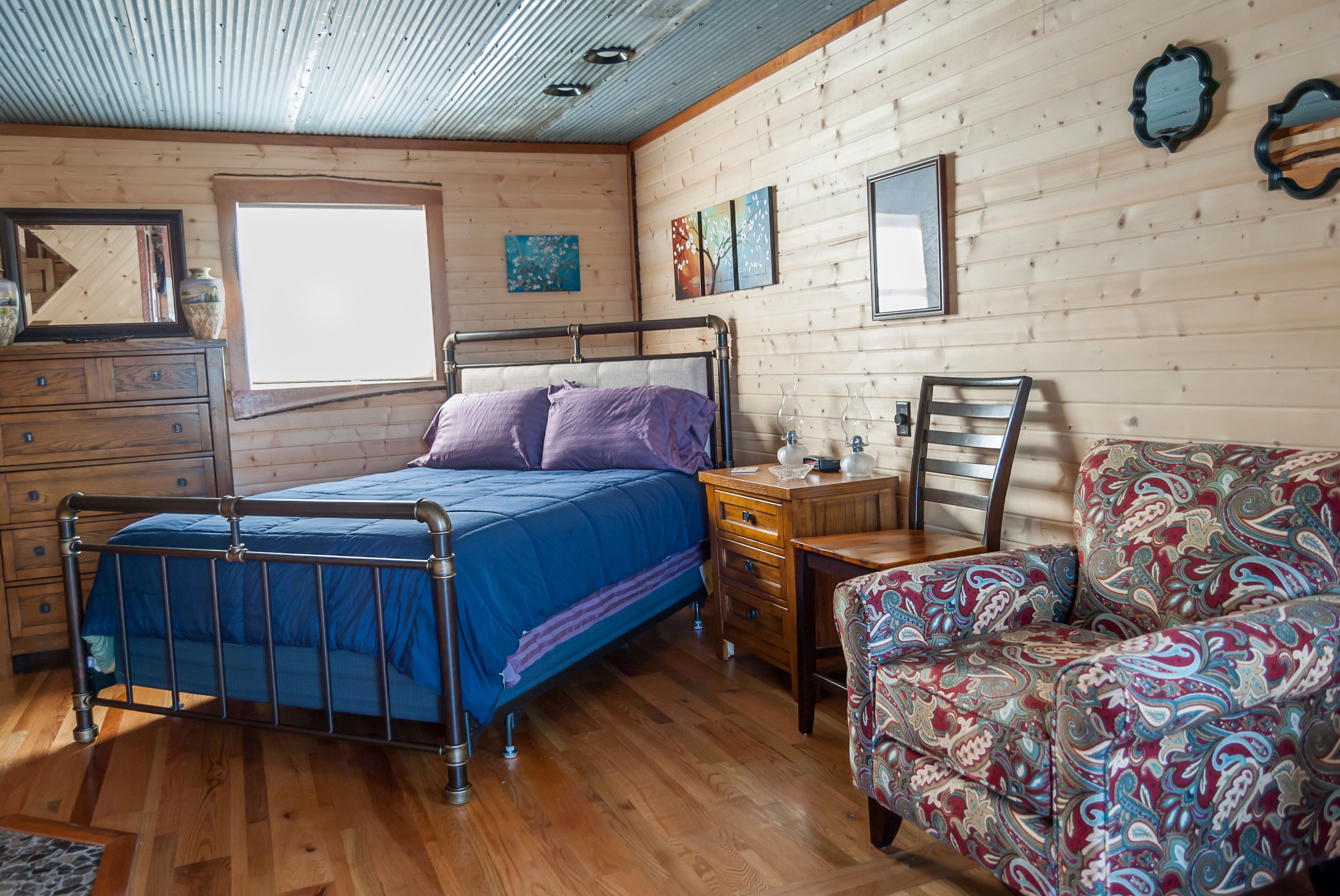 A bedroom with a metal bed frame, a wooden dresser, a patterned armchair, and a window with artwork on the wall.