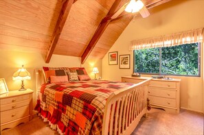 A cozy bedroom with a wooden ceiling, a large bed with a quilt, bedside tables with lamps, a window with a view of trees, and framed pictures on the wall. - ROMANTIC LOG CABIN ~ Quiet, Clean, Cozy ~Walk to Trails, 3 Min to Town! (Idyllwild-Pine Cove)