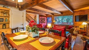 A cozy log cabin interior with a dining table set for four, a red leather sofa, and a staircase leading to an upper level.