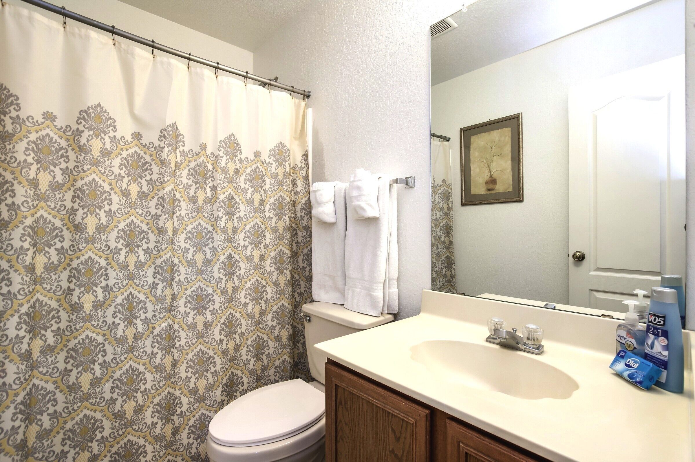 Un baño con cortina de ducha estampada, inodoro, lavamanos con artículos de aseo y espejo.