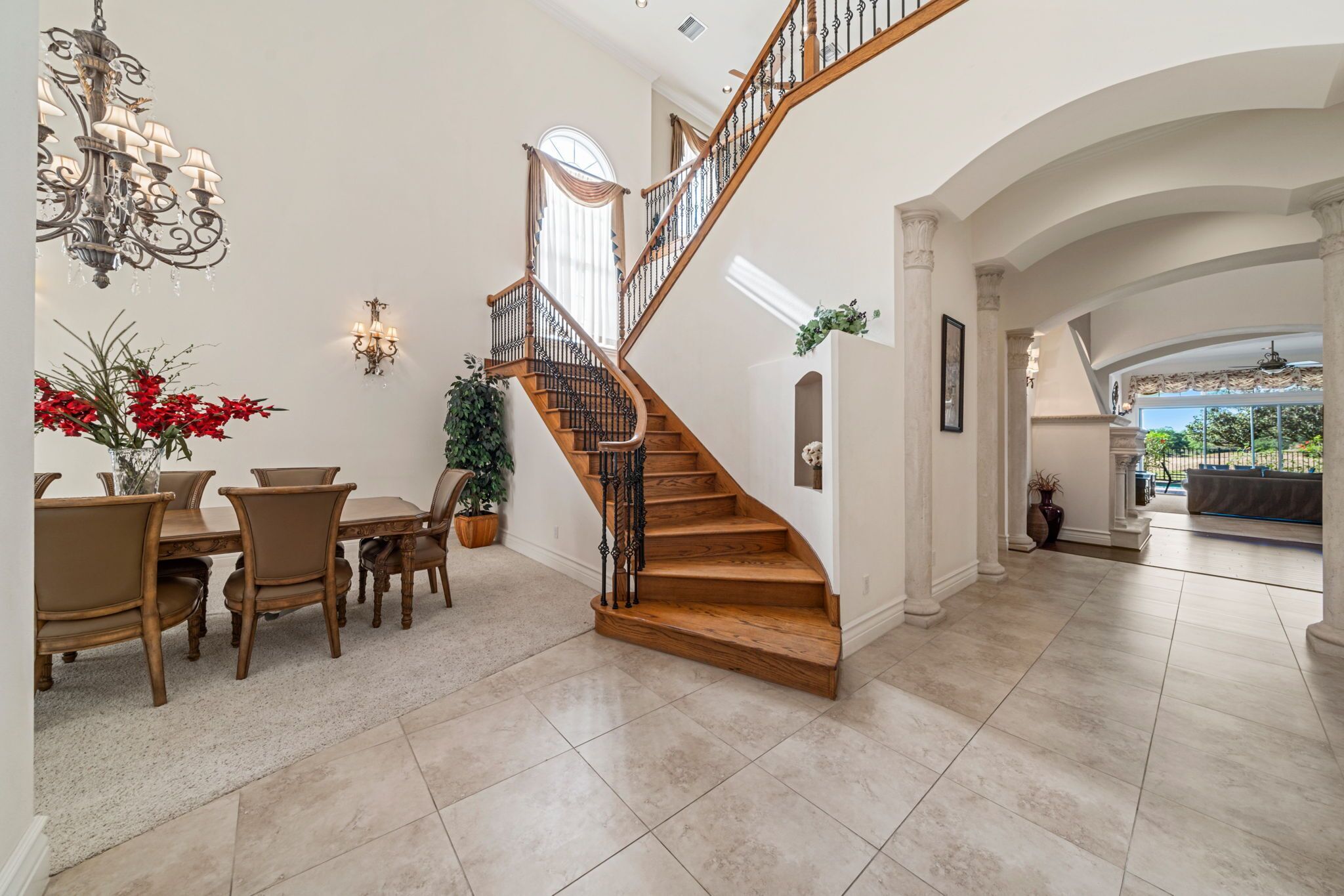 A spacious interior with a wooden staircase, a dining area with chairs and a table, and a view of the outdoors through a large window.