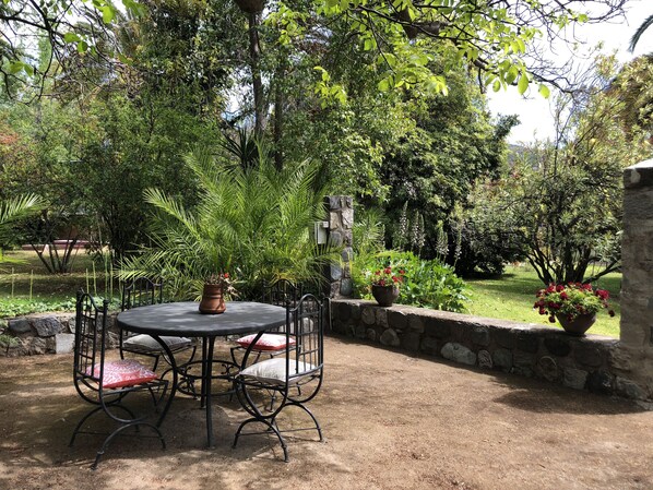 Une table ronde entourée de chaises et agrémentée d’une plante en pot, le tout situé dans un espace jardinier bordé de murs en pierre et orné de diverses plantes.