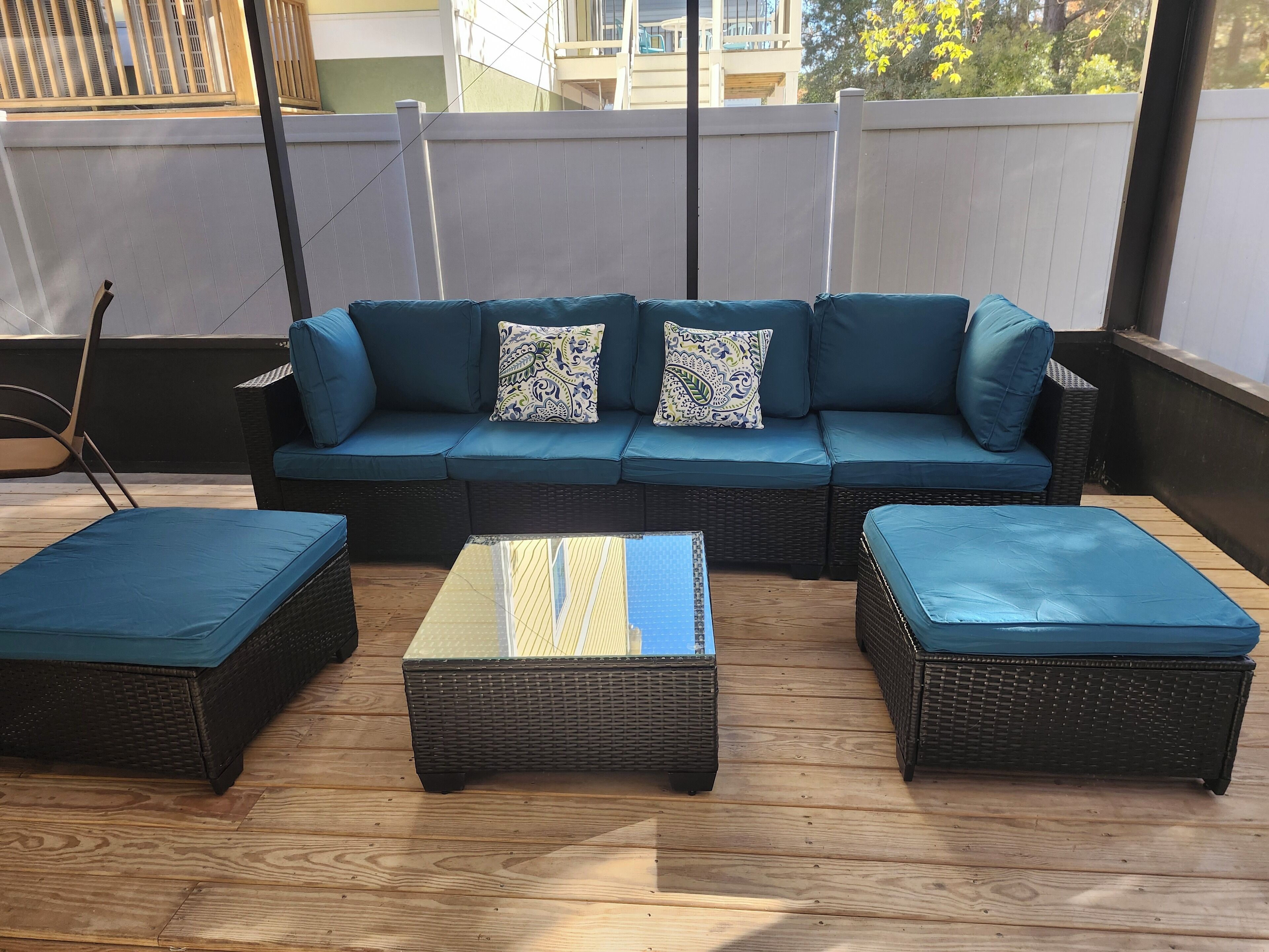 A covered outdoor seating area with a dark wicker sofa and matching ottomans, a wooden floor, and a white fence in the background.