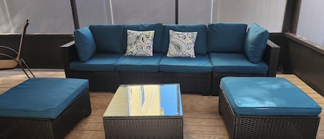 A covered outdoor seating area with a dark wicker sofa and matching ottomans, a wooden floor, and a white fence in the background.