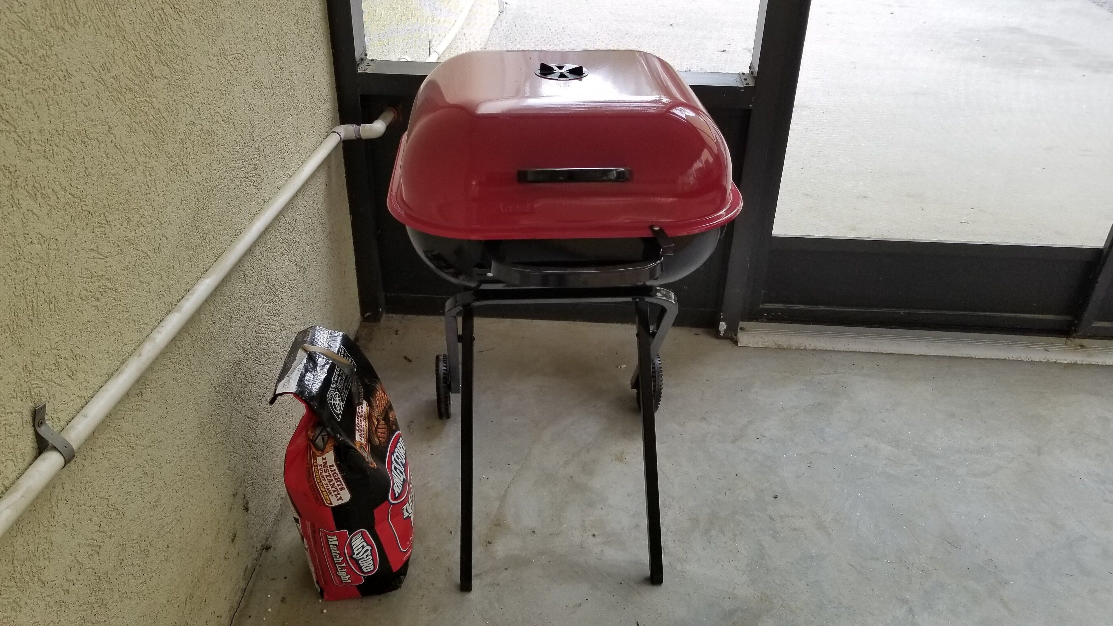 Una parrilla roja con un soporte negro, ubicada junto a una pared color beige y una ventana.