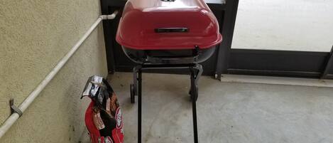 Una parrilla roja con un soporte negro, ubicada junto a una pared color beige y una ventana.