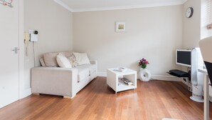 A modern living room with a sofa, a small white side table, a white vase with flowers, a flat-screen TV, and a ceiling-mounted fan. - Cozy Fitzrovia studio on a quiet cobbled street in the West End! (London)