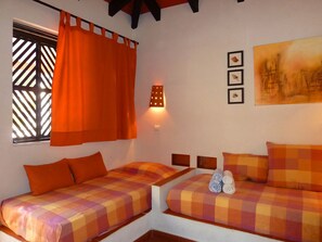 A room with two beds, each with orange bedding and a checkered pattern. There is a window with a lattice design and orange curtains. A wall-mounted light fixture is visible, and three framed pictures are hung on the wall.