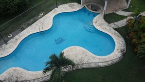 En bild tagen från luften som visar en pool med en böjd form, omgiven av en betongplattform och en liten paviljong med ett tak täckt av kakel.