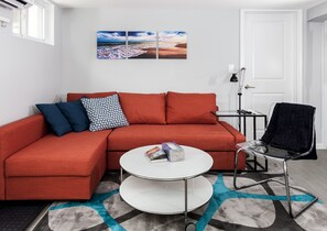 Un salon moderne avec un canapé rouge, une table basse en verre et un fauteuil noir. Un tableau représentant une scène de plage est accroché au mur.