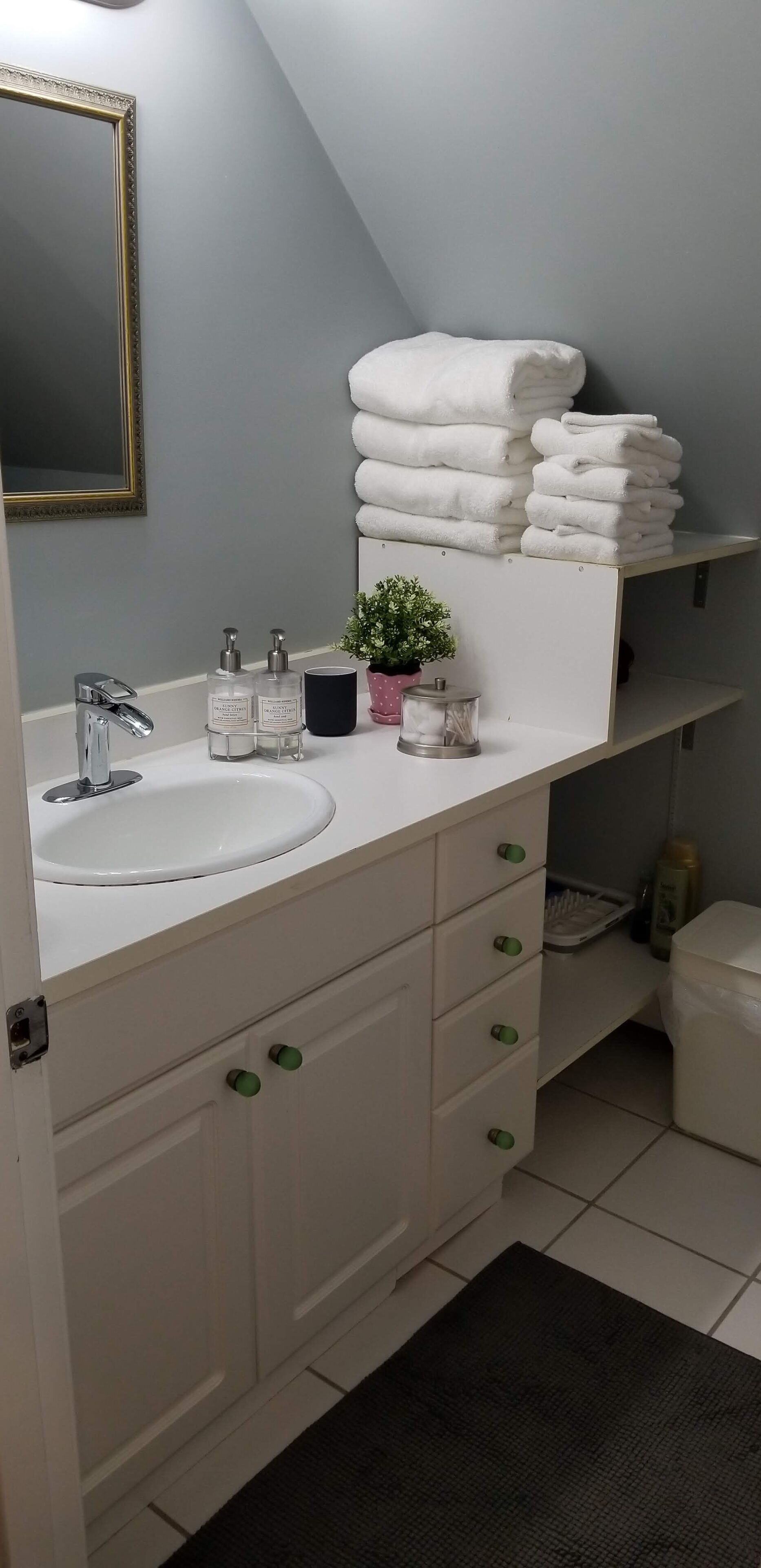 A bathroom with a white sink, a mirror with a gold frame, a stack of white towels, a small potted plant, and toiletries on the counter.
