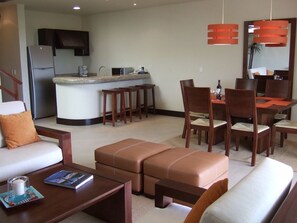 A modern living room with a sofa, ottoman, coffee table, and dining area with chairs and a table. A kitchen with a refrigerator and bar stools is visible in the background.