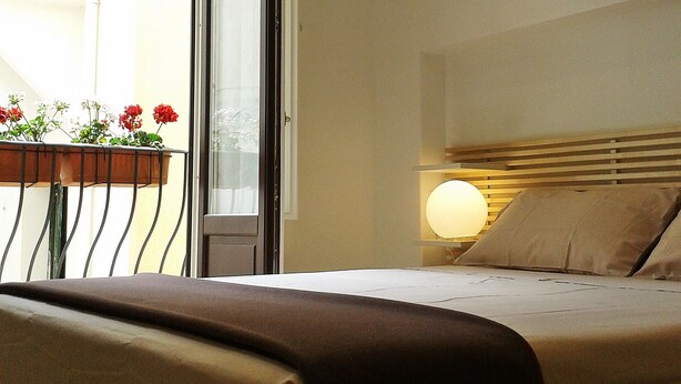 Ein Schlafzimmer mit Bett, Nachttischlampe, Fenster mit Blick auf einen Balkon mit Blumen und einer zur Außenseite führenden Tür.