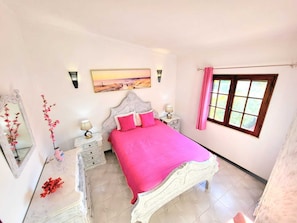 A bedroom with a large bed, a nightstand, a dresser, a mirror, a window with curtains, and a painting on the wall.