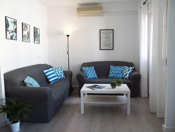 A living room with two grey sofas, a white coffee table, and a floor lamp. There are framed pictures on the wall and a potted plant on the floor.