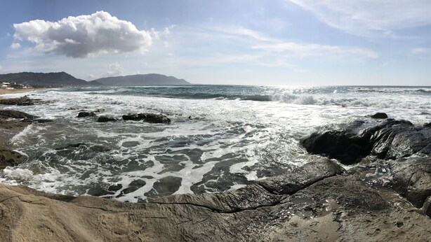 Une plage rocheuse où les vagues se brisent sur le rivage, un ciel dégagé et des montagnes à l’horizon.