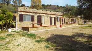 Un bâtiment en pierre doté de nombreuses fenêtres et de volets, entouré d’une zone aride et herbeuse où la végétation est clairsemée.
