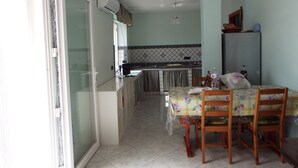 Un coin repas avec une table et des chaises, une cuisine en arrière-plan et une porte-fenêtre coulissante donnant sur un espace extérieur.