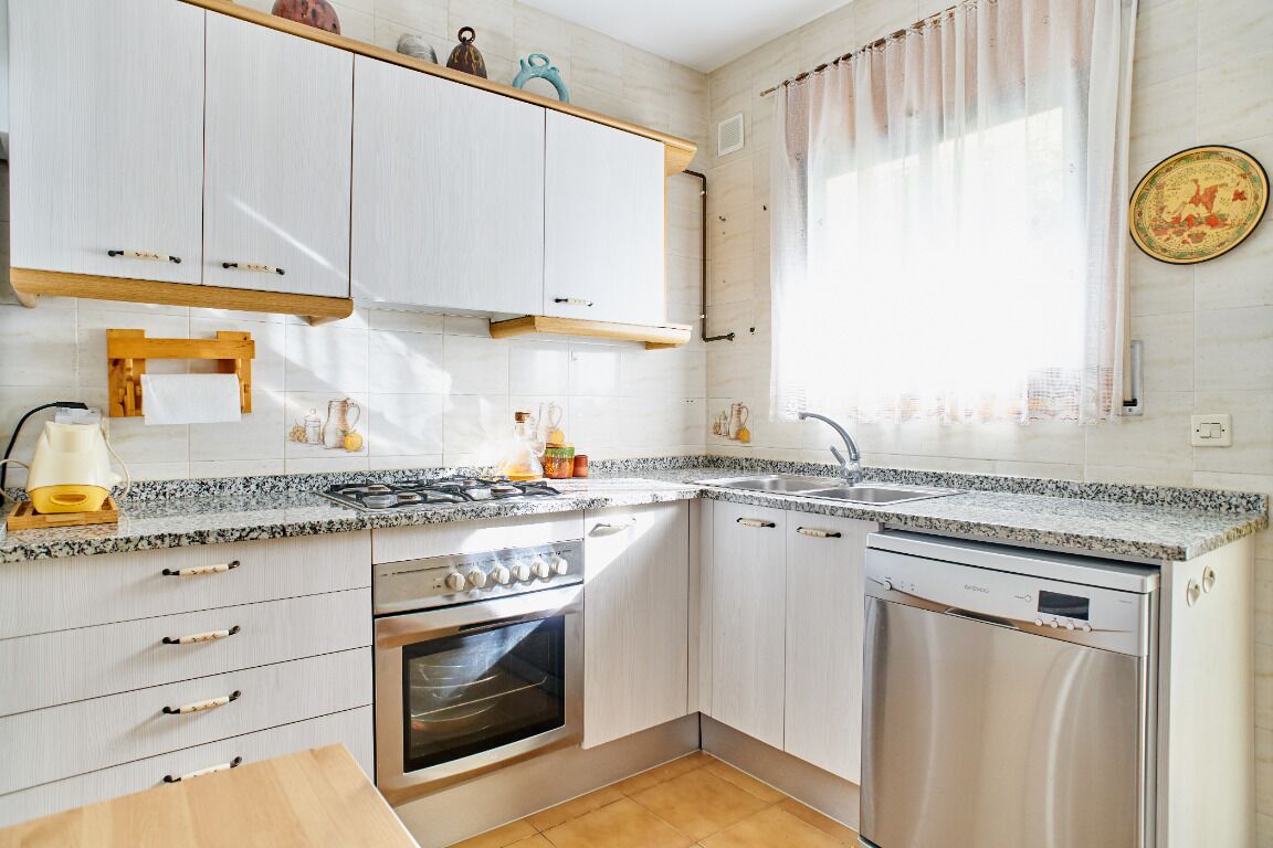 Una cocina moderna con gabinetes blancos, encimera de granito, horno integrado, lavavajillas y una ventana con cortinas.