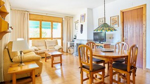A cozy living room with a sofa, a coffee table, and a dining area with a wooden table and chairs. There is a TV on a stand, a lamp, and a window with curtains.