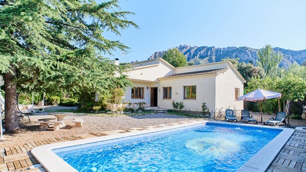 A swimming pool with a wooden deck, a white building with a tiled roof, and a mountainous backdrop.
