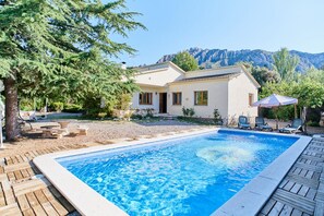 Una piscina con cubierta de madera, un edificio blanco con techo de tejas y montañas al fondo.