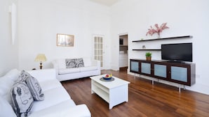 Un salon moderne avec un canapé blanc, une table basse blanche, un téléviseur posé sur un meuble et une porte donnant sur la cuisine.