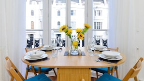 Une table à manger dressée pour deux personnes, avec des chaises rembourrées bleues, un vase rempli de tournesols et une vue sur les bâtiments à travers de grandes fenêtres.