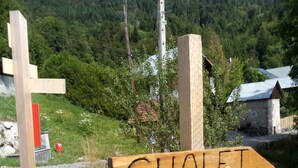 Et træskilt med et hjortehoved og teksten "CHALET des CERES", der peger mod højre, foran et bakket landskab med grønt og en mindre bygning.