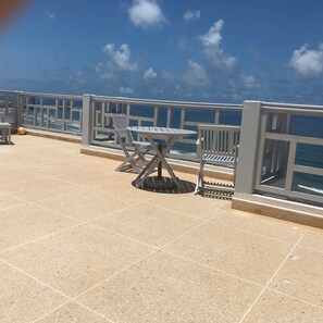 A tiled outdoor area with a table and chairs, a railing, and a view of the sea.