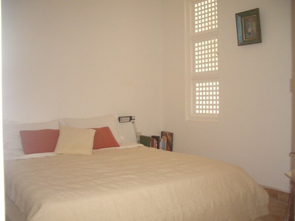 A bedroom with a bed, pillows, a bedside table with books, a lamp, and a window with blinds.