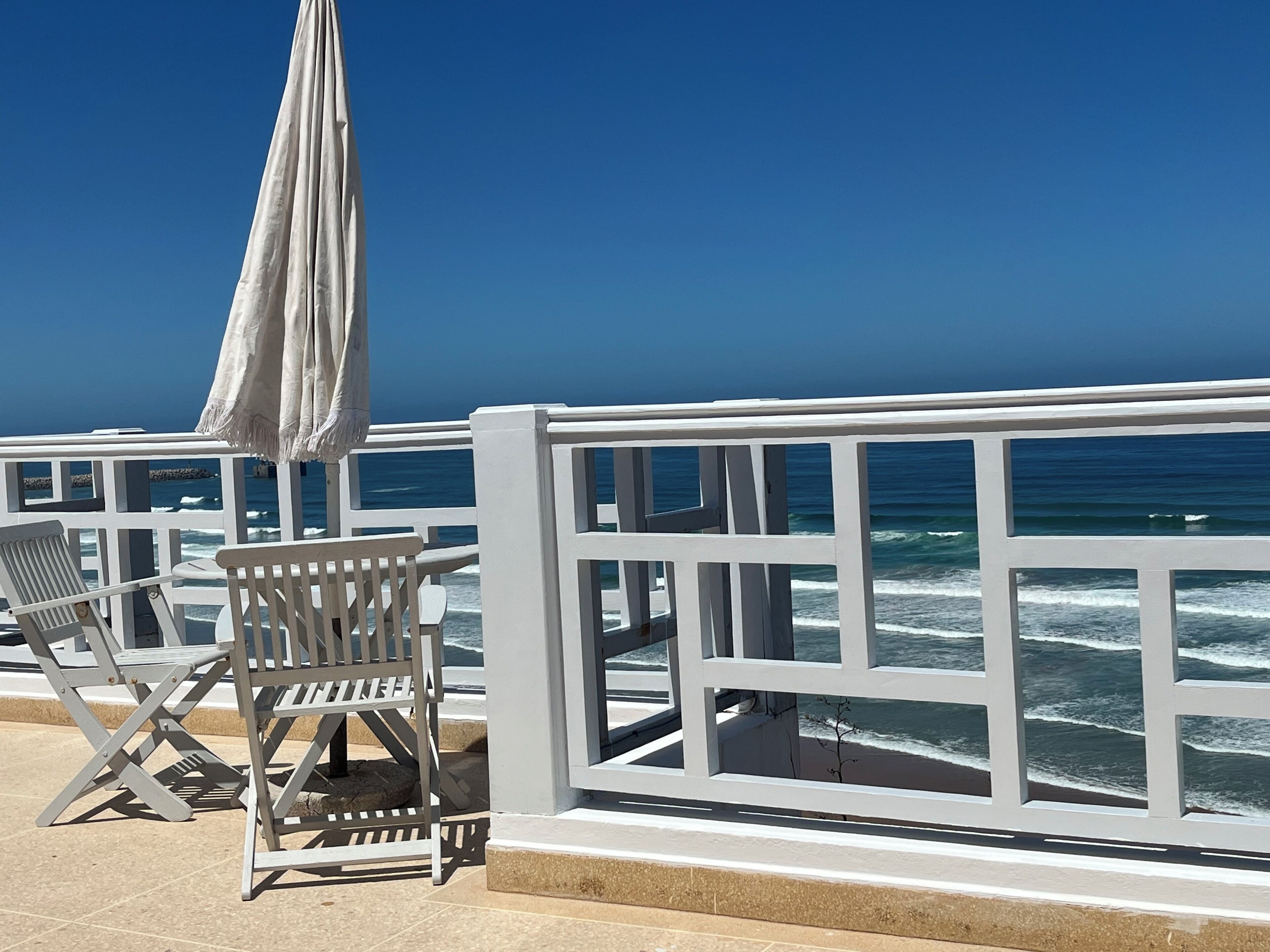 A seaside terrace with white railings, a white umbrella, and a chair.