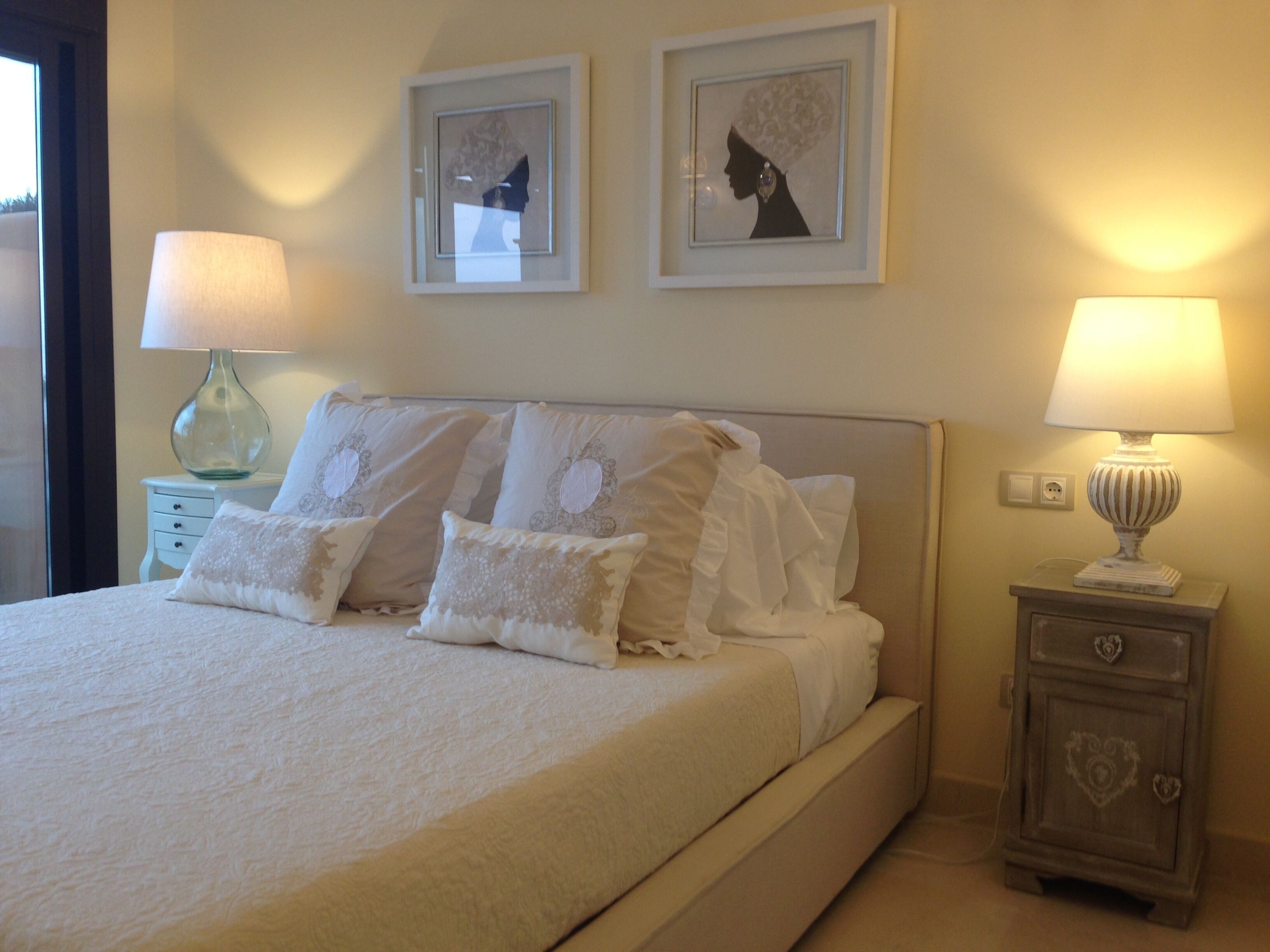 Een net opgemaakt bed met witte lakens en decoratieve kussens, een nachtkastje met een lamp, en twee ingelijste kunstwerken aan de muur.