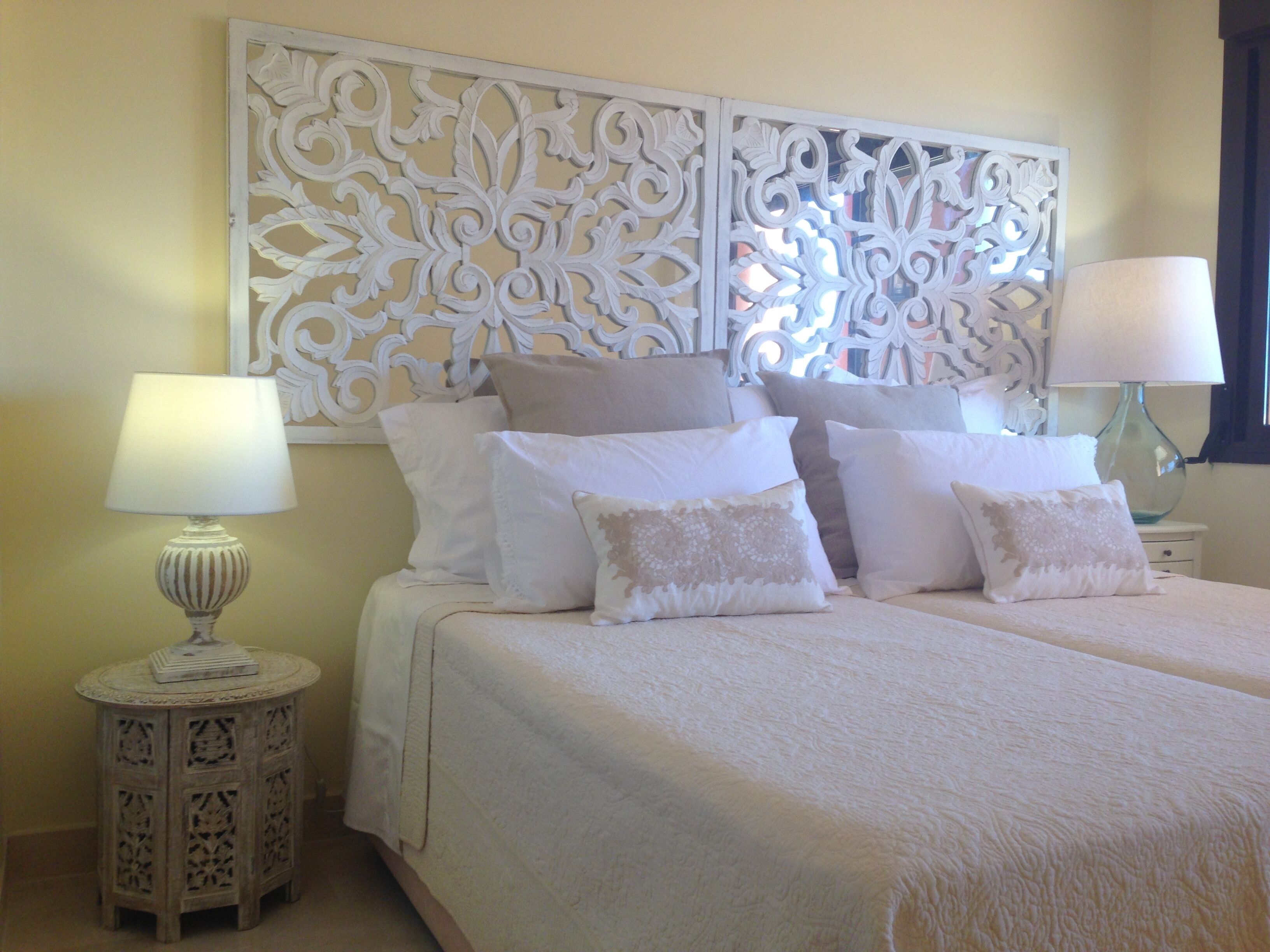 A double bed with a white bedspread and multiple pillows, a decorative white headboard, a bedside lamp, and a small round side table.