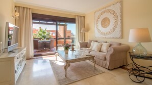 Un salon avec un canapé, une table basse et un téléviseur. Il y a un balcon d’où l’on peut admirer des immeubles et une plante.