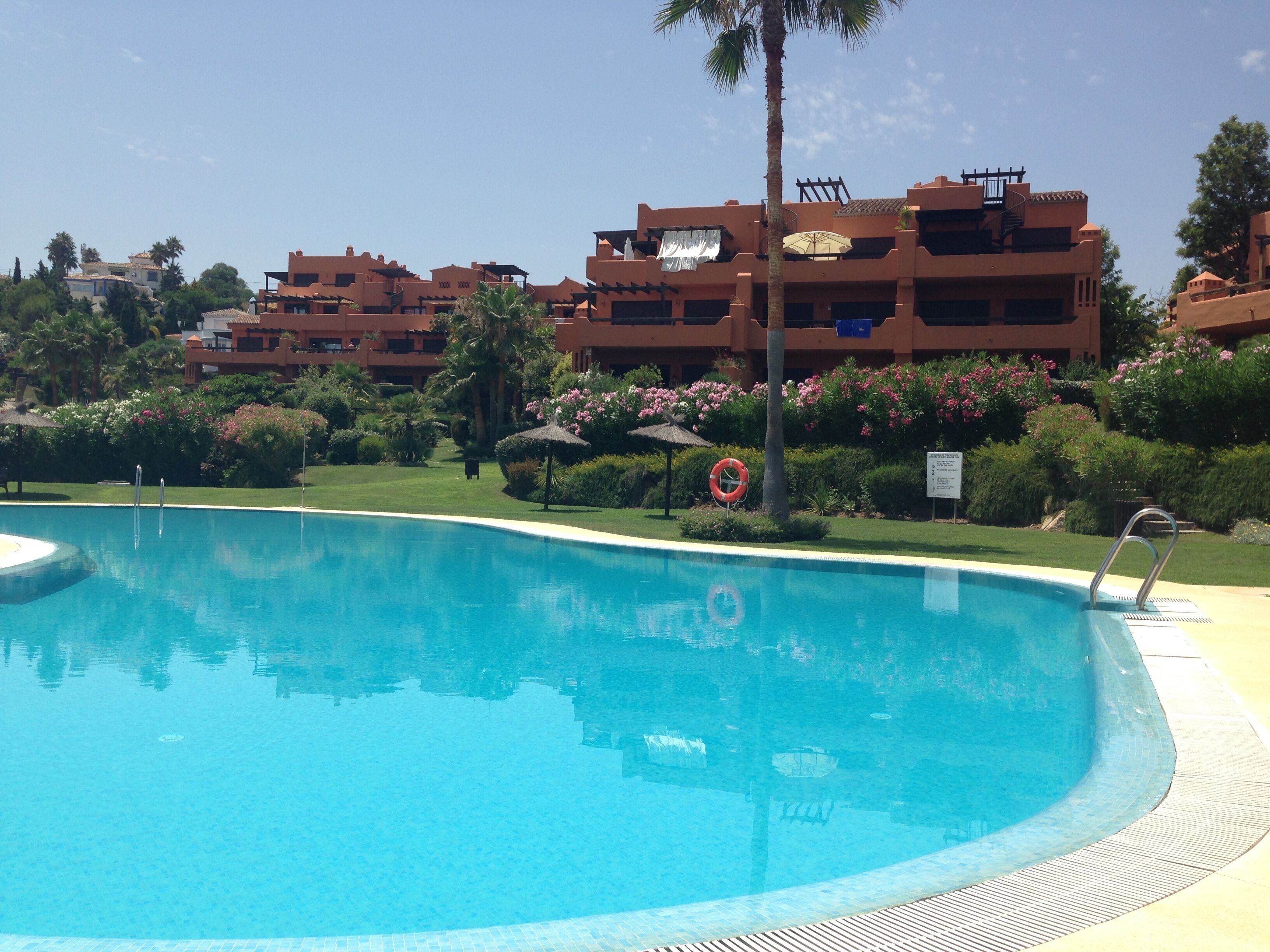 En swimmingpool med en klar, blå vandoverflade, omgivet af en velplejet græsplæne og diverse planter. I baggrunden ses etageejendomme med altaner, sandsynligvis en del af et hotel eller resort.