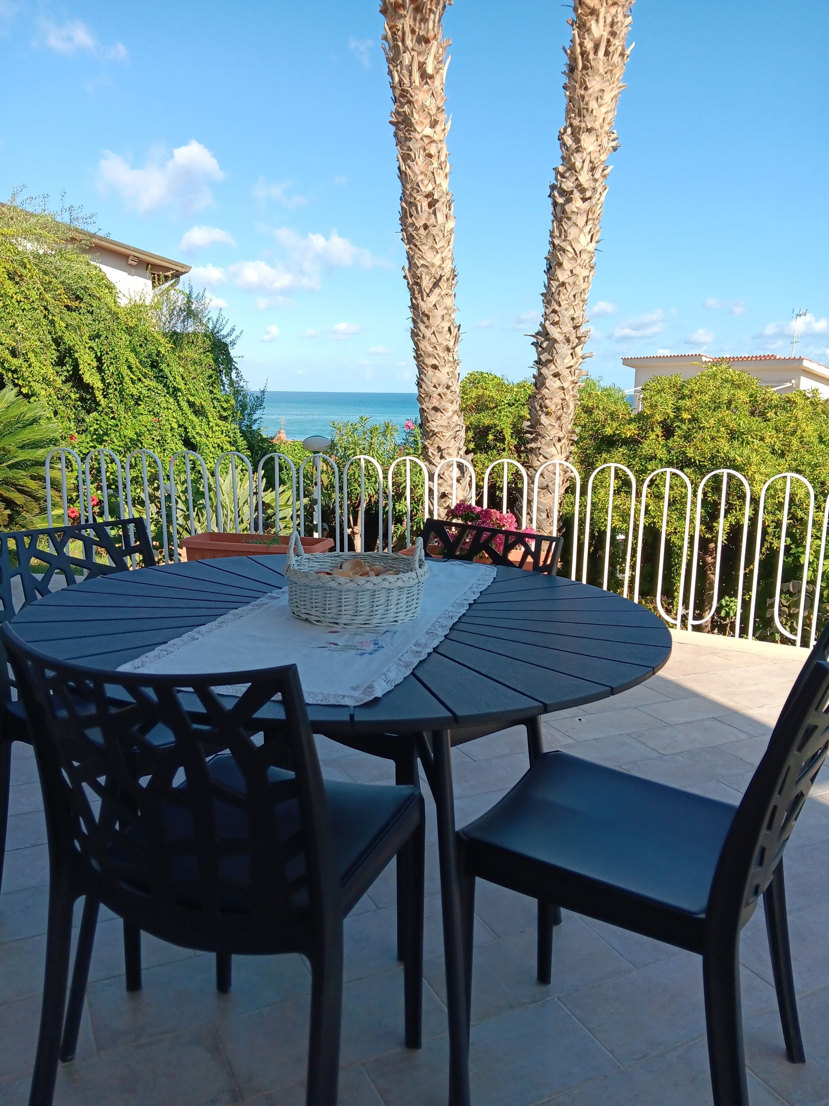 An outdoor patio with a round table and chairs, a metal fence, and a view of the sea.