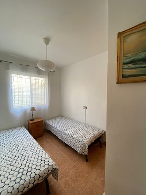 Una habitación pequeña y limpia con dos camas individuales, una mesita de noche de madera, una lámpara colgante y una ventana con barrotes decorativos.