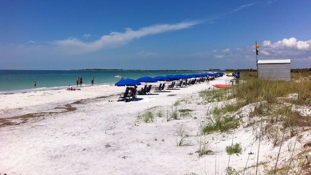 Una playa con arena blanca, sombrillas azules y un pequeño edificio con una bandera.