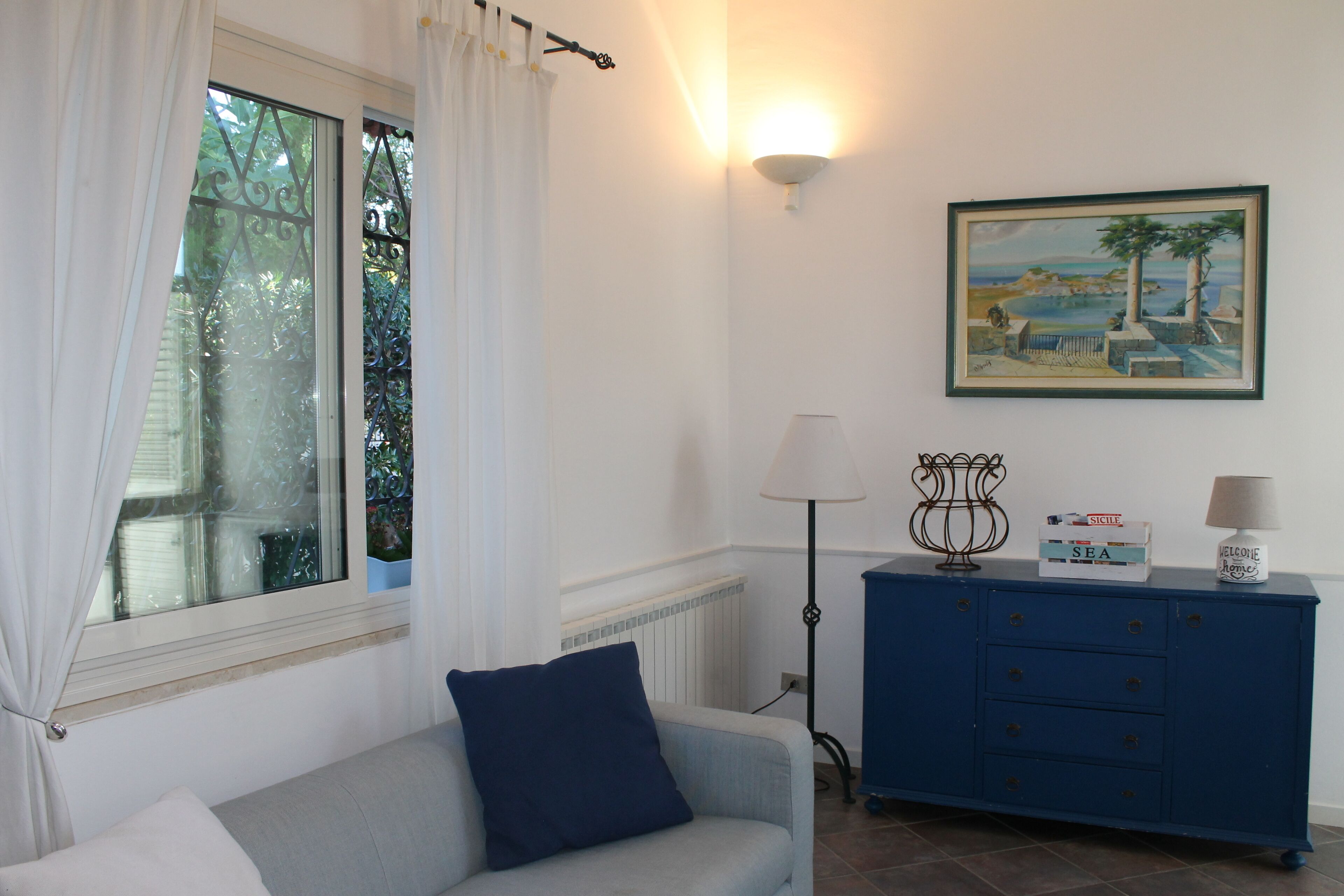 A room with a window, a blue cabinet, a lamp, and a framed painting on the wall.