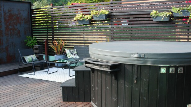 Een hottub op een houten terras, met buitenmeubilair en plantenbakken tegen een houten hek.